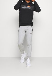 Sweat-shirt noir à capuche avec logo gris et pantalon de survêtement gris. Le pantalon de survêtement est doté de poches latérales zippées. Le mannequin porte des baskets blanches.