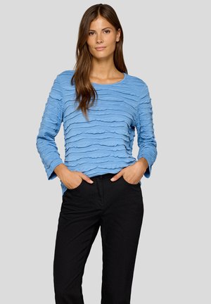 Hellblaues Langarmshirt mit strukturiertem, geschichtetem Stoffdesign. Getragen mit schwarzen Hosen, rundem Ausschnitt und lockerer Passform.