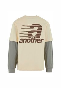 T-shirt crème avec des manches longues grises contrastées. Présente un design graphique audacieux en brun avec du texte et des lignes au dos.