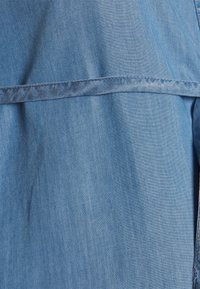 Tecido de denim numa tonalidade azul média com uma textura suave. Apresenta uma costura horizontal ao longo do material, indicando um design em camadas.