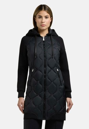 Młoda kobieta ubrana w czarną pikowaną, długą bluzę z kapturem, z przednim zamkiem błyskawicznym i zapinanymi na zamek kieszeniami, stojąca na jednolitym tle.