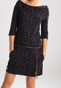 Svart klänning med båtöppning, trekvartsärmar och flerfärgat prickmönster. Har sidofickor och ett flätat bälte.