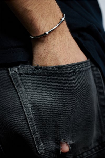 Bracciale in metallo argentato con un design liscio e arrotondato, caratterizzato da estremità affusolate. Indossato su un polso, parzialmente visibile dietro a dei jeans neri usurati.