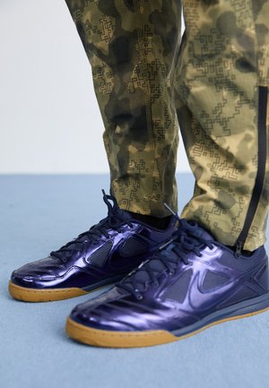 Purple athletic shoes with glossy finish, combined materials, and gum rubber sole; paired with green camo patterned pants featuring zippers.