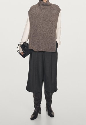 Person trägt eine braune, gestrickte ärmellose Rollkragenweste über einem weißen Langarmshirt, schwarze Hosen mit weitem Bein, schwarze Stiefel und hält eine schwarze Handtasche.