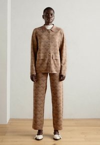 Beige gemusteter Anzug mit einem kragenlosen Jacket mit Knopfverschluss und zwei Taschen, kombiniert mit gerade geschnittenen Hosen. Die Schuhe sind weiß.