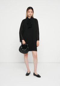 Μαύρο, πλισέ φόρεμα με μακριά μανίκια και λεπτομέρεια με κορδέλα στο λαιμό. Συνδυασμένο με μια μαύρη, ομαλή τσάντα και επίπεδα μαύρα παπούτσια.