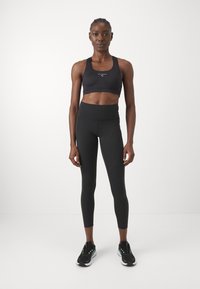 Soutien-gorge de sport noir avec logo et leggings noirs taille haute. Le modèle porte des chaussures de sport noires avec des accents verts. Tissu lisse et extensible.