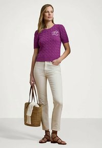 Γυναίκα που φοράει ένα μωβ κοντομάνικο πουλόβερ με πλέξη, λευκά τζιν, καφέ σανδάλια και κρατάει μια πλεκτή τσάντα με λευκό πανί.