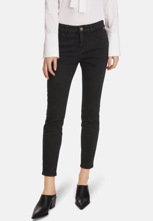 Kvinde iført slanke sorte jeans, hvid bluse med lange ærmer og sorte spidse hælede mules mod en hvid baggrund.