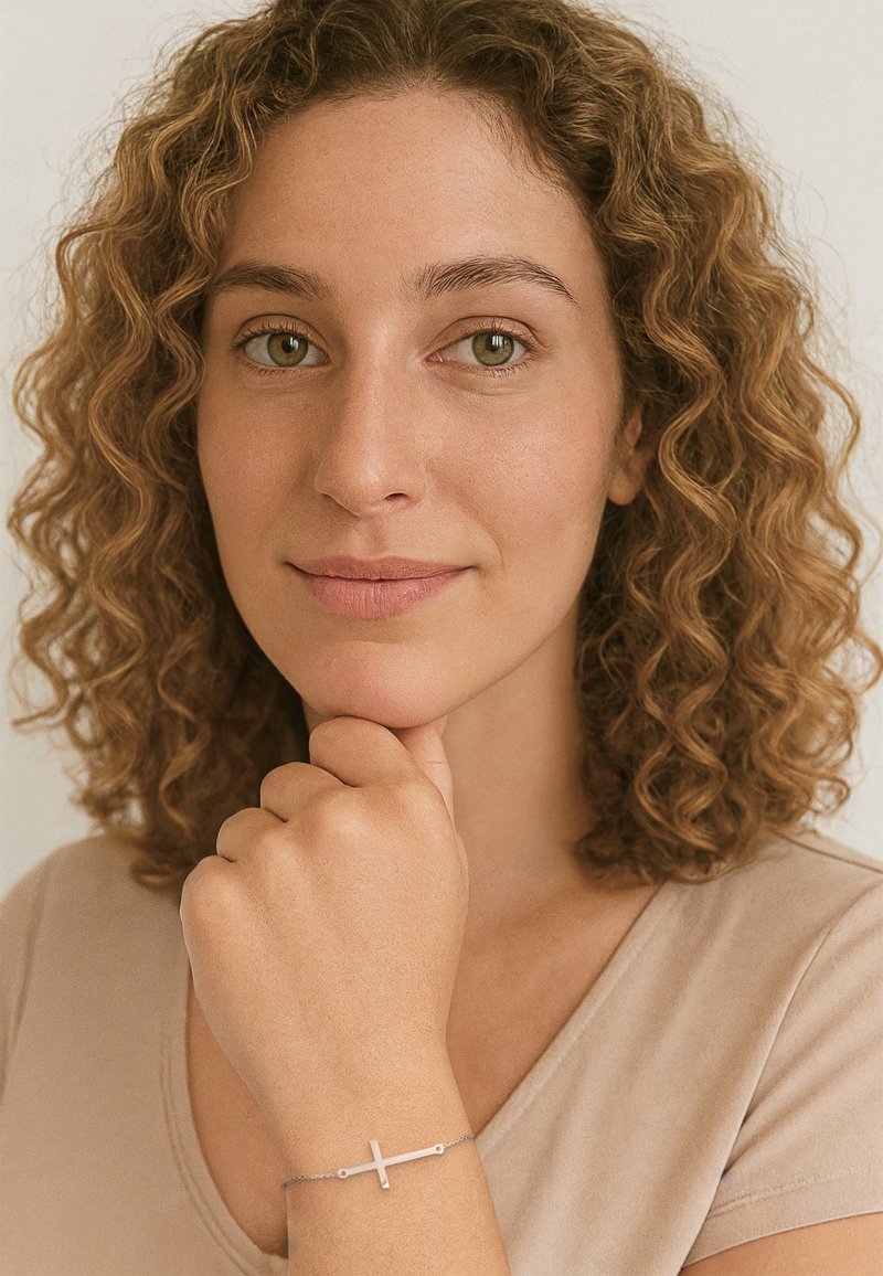 Bracciale d'argento con un design a croce orizzontale, caratterizzato da una delicata catena e una superficie liscia, indossato su una mano che riposa vicino al mento.