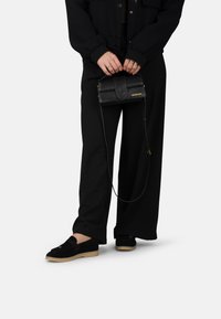 Zwarte suède schoenen met een lichtbruine zool, losse zwarte broek en een kleine zwarte leren schoudertas met een gouden logo en schouderband.