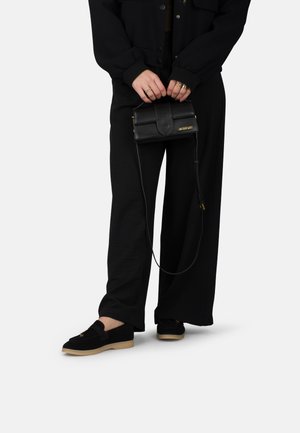 Zwarte suède schoenen met een lichtbruine zool, losse zwarte broek en een kleine zwarte leren schoudertas met een gouden logo en schouderband.