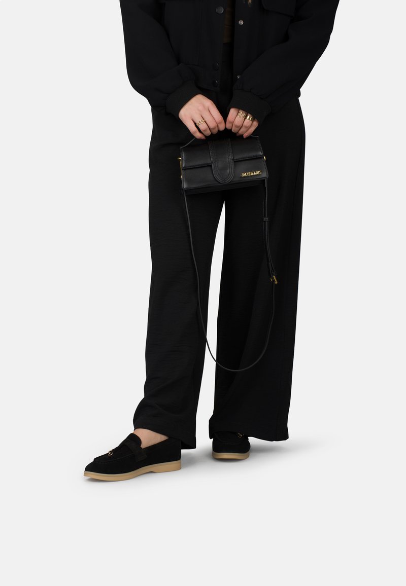 Zwarte suède schoenen met een lichtbruine zool, losse zwarte broek en een kleine zwarte leren schoudertas met een gouden logo en schouderband.