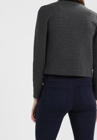 Šedá ribbovaná krátká bunda s kulatým výstřihem, v kombinaci s tmavě modrými džíny. Tkanina má texturovaný vzhled a strukturovaný střih.