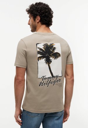 Homme portant un t-shirt beige avec un motif de palmier noir et le texte "Tommy Hilfiger" dans le dos, associé à un jean bleu.