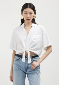 Donna con capelli neri lisci, che indossa grandi orecchini d'oro, una maglietta bianca a maniche corte annodata in vita, jeans azzurri chiari e una cintura nera.