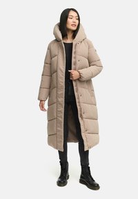 Lange pufferjas in lichtbeige met een afneembare capuchon, doorgestikte textuur, knoopsluiting aan de voorkant en zijzakken, gecombineerd met zwarte jeans en laarzen.