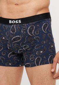 Marineblaue Boxershorts mit einem Paisleymuster in Grautönen und Weiß, ausgestattet mit einem schwarzen Bund, der das "BOSS"-Logo in Weiß trägt.