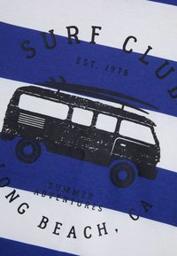 Grafiska T-kreklu ar ziliem un baltiem horizontāliem svītrām. Melna druka ar vintage furgonu, kas pārvadā sērfa dēļus, un teksts "Sērfa klubs" ar atrašanās vietas detaļām.