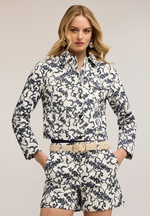 Kobieta o blond włosach, ubrana w biały komplet z kurtką i szortami z abstrakcyjnym czarnym wzorem, beżowy pleciony pasek, kolczyki, ręce w kieszeniach.