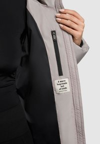 Světle šedá prošívaná bunda uvnitř s černou textilní podšívkou, s kapsou na zip a štítkem, který říká "Teplé objetí vyrobené z látky."