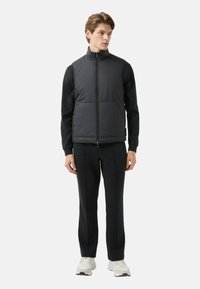 Czarna pikowana kamizelka z wysokim kołnierzem, zapięciem na zamek i dwoma przednimi kieszeniami, noszona na czarnej bluzce z długim rękawem, zestawiona z ciemnymi spodniej dresowymi.