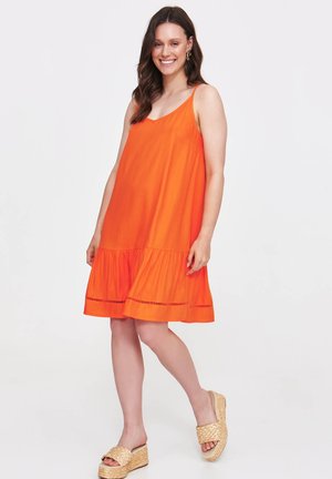 Pomarańczowa sukienka bez rękawów z falbaną na dole, uszyta z lekkiego materiału. Modelka łączy ją z plecionymi sandałami na platformie.