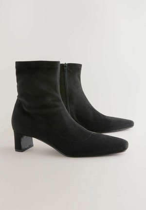 Černé semišové kotníkové boty s špičatou špičkou, zipovým uzavřením a nízkým blokovým podpatkem se lesklým černým akcentem. Hladká textura a elegantní design.