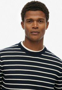 Granatowy i biały sweter z paskami, z ryflowanym okrągłym dekoltem. Miękka tekstura, dopasowany krój oraz równomierny wzór pasków na całej powierzchni.