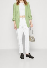 Ljusgrön blazer över en vit kort topp, matchad med vita löst sittande byxor och svarta klackskor; håller en pärlprydd silverhandväska.