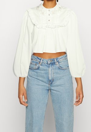Mujer con blusa blanca corta de manga larga con cuello con volantes y vaqueros de talle alto de color azul claro, de pie frente a un fondo liso.