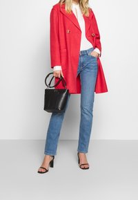Abrigo de mezcla de lana rojo con diseño de doble botonadura, botones dorados, combinado con jeans de color azul claro y un bolso de mano de cuero negro. Sandalias de tacón negras.