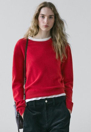 Ung kvinde med langt bølget blondt hår iført en rød sweater, hvid undertrøje, mørke bukser og en sort skuldertaske mod en ensfarvet baggrund.