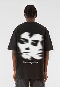 Sort bomulds T-shirt med et sløret grafik af ansigtter bagpå, med hvid tekst nedenunder, korte ærmer og en afslappet pasform.