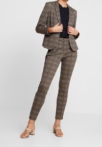 Costume sur mesure à carreaux dans des tons de brun avec une veste cintrée et un pantalon slim. Porté avec un haut noir et des sandales à talons blocs beiges.