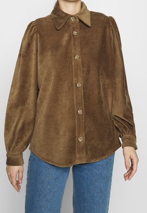 Brązowa koszula sztruksowa zapinana na guziki z bufkami na długich rękawach, zestawiona z niebieskimi dżinsami, prezentowana na stojącej osobie.