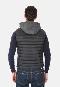 Uomo che indossa un gilet trapuntato nero con cappuccio grigio su una maglietta a maniche lunghe blu navy e jeans blu, in piedi di spalle su uno sfondo bianco.