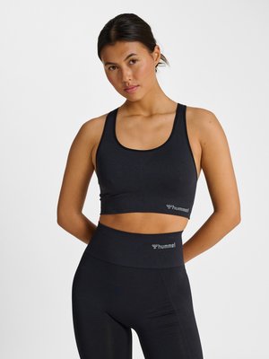 Ung kvinde i sort atletisk crop top og højtaljede leggings står med hænderne bag ryggen mod en ensfarvet hvid baggrund.
