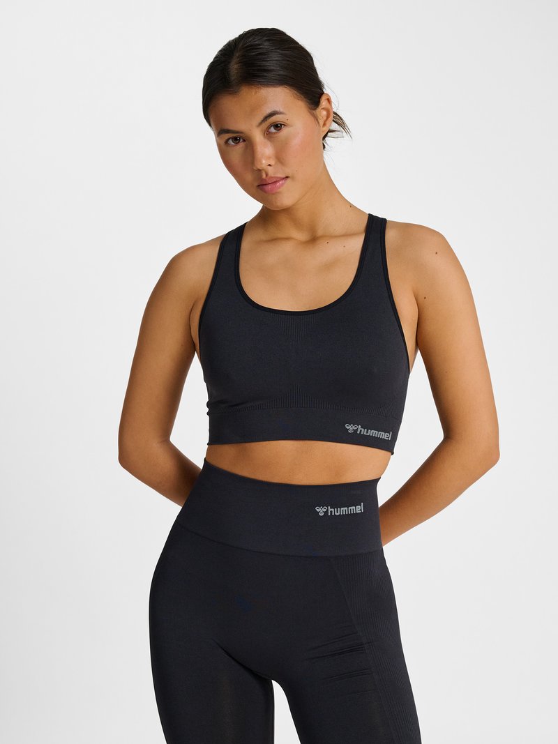 Joven mujer con top corto deportivo negro y leggings de cintura alta, de pie con las manos detrás de la espalda contra un fondo blanco liso.
