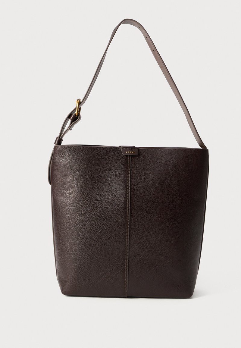 Bolso de hombro de cuero marrón oscuro con correa ajustable, hebilla dorada y pequeño detalle de logo central sobre un fondo liso.