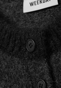 Zwarte gebreide cardigan met een gestructureerd oppervlak, voorzien van ronde zwarte knopen en een stoffen label met de tekst "WEEKDAY."