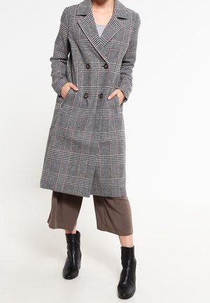 Personne portant un long manteau à carreaux noir et blanc à double boutonnage, un pantalon marron à jambes larges et des bottines noires. Les mains sont dans les poches du manteau.
