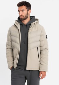 Giacca puffer beige chiaro con cappuccio, caratterizzata da pannelli trapuntati, dettagli in zip neri e una texture liscia, indossata sopra una felpa grigia.