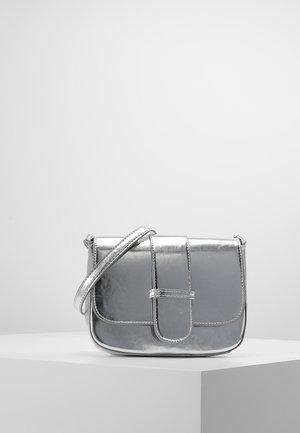 Malá stříbrná kovová crossbody taška s klopou a popruhem vystavená na bílém podstavci před šedým pozadím.