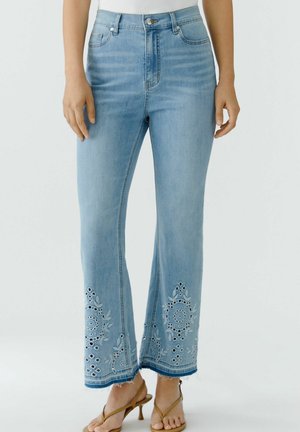 Jean bleu clair taille haute à jambes larges, broderie florale en découpe près des ourlets effilochés, porté avec des sandales à talons couleur fauve.