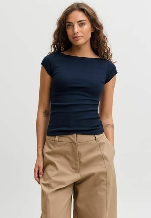Jeune femme aux cheveux bouclés portant un haut bleu marine ajusté à manches courtes et un pantalon beige à jambes larges, debout devant un fond uni.