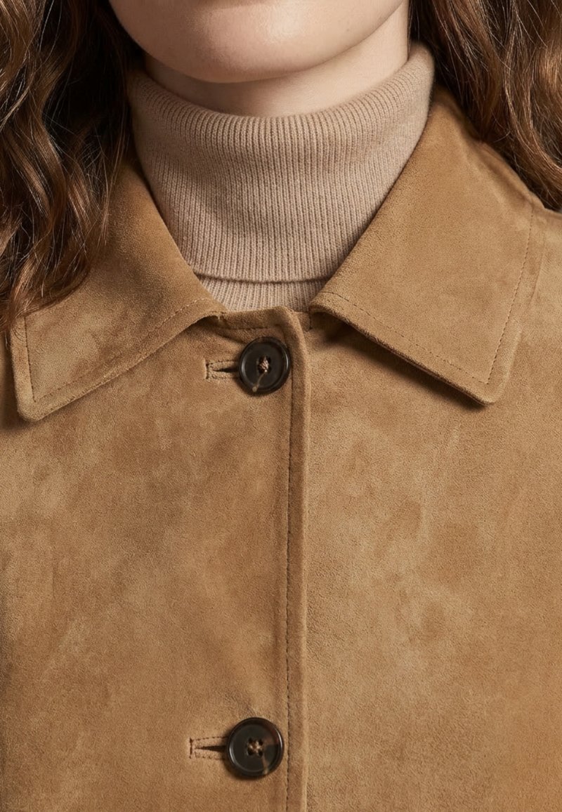 Femme portant un manteau en suède beige avec de gros boutons par-dessus un pull à col roulé côtelé marron clair, cheveux partiellement visibles sur les épaules.