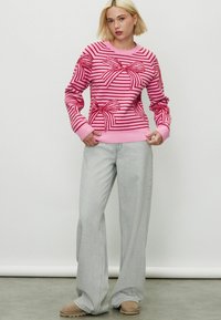 Pull en rayures roses avec des motifs de nœuds rouges, décolleté rond et manches longues. Associé à un jean large gris clair et des chaussures beiges.