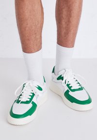 Baskets avec une base blanche et des accents verts, présentant une texture perforée, un design à lacets, et une semelle épaisse et texturée. Chaussettes blanches visibles.